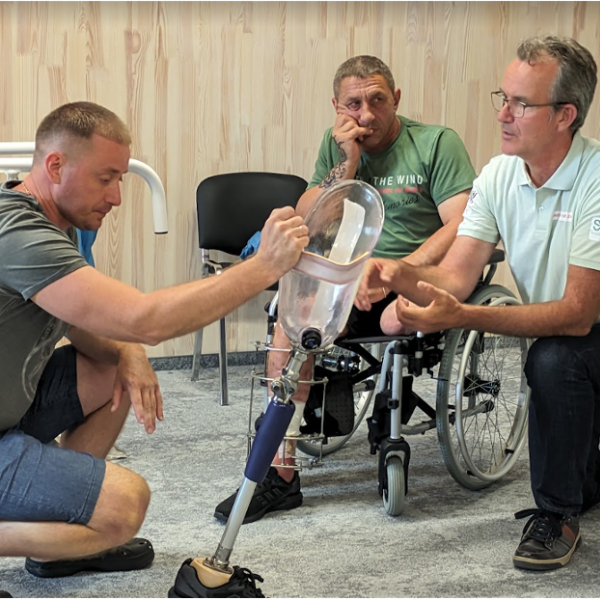 Three man in the picture, one to the left kneeling and holding a prosthesis on the floor, one to the right sitting on a chair and one in the middle sitting in a wheelchair